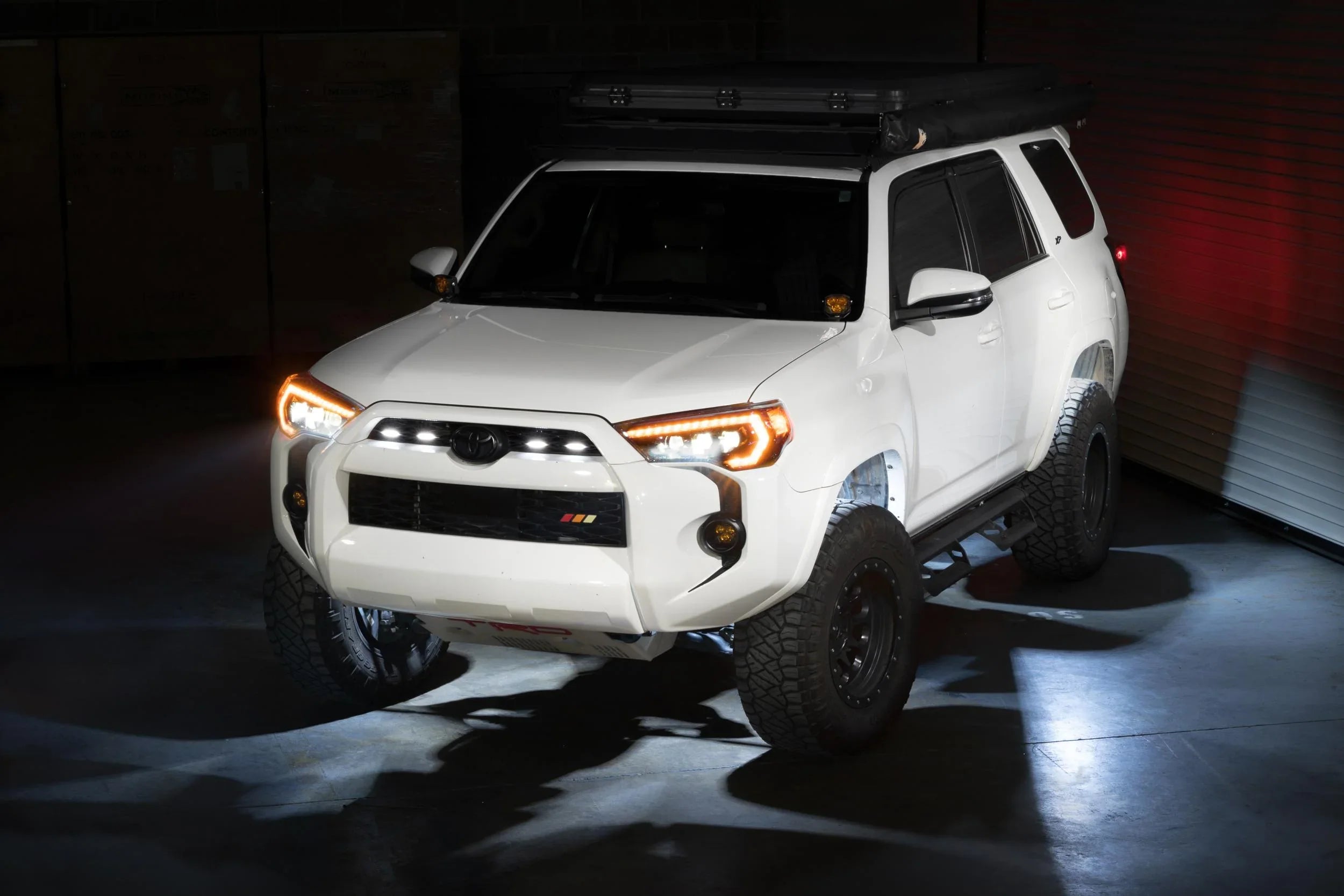 White Toyota 4Runner with off-road tires, roof rack, and LED lights in a dim garage.