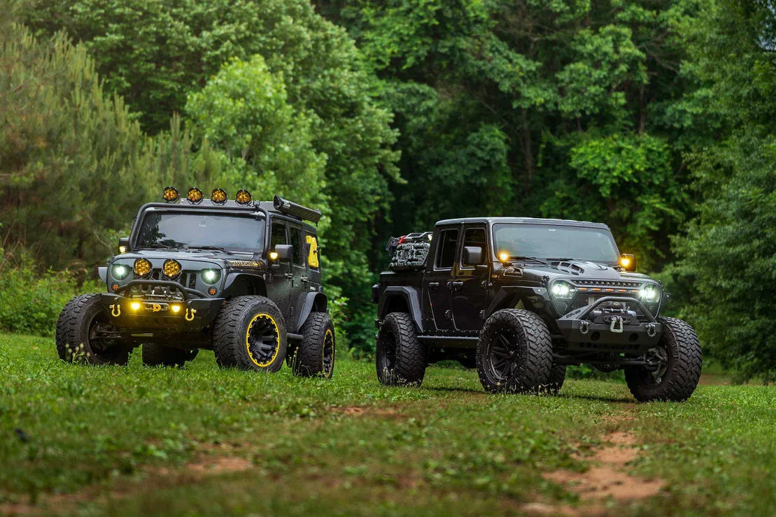Two modified Jeep Wranglers with off-road gear and large tires parked in a forest clearing.