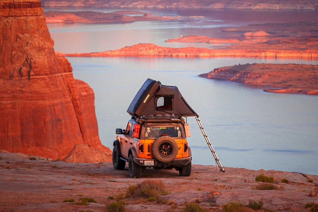 Orange off-road SUV with roof top tent and ladder camping by a scenic canyon lake at sunset