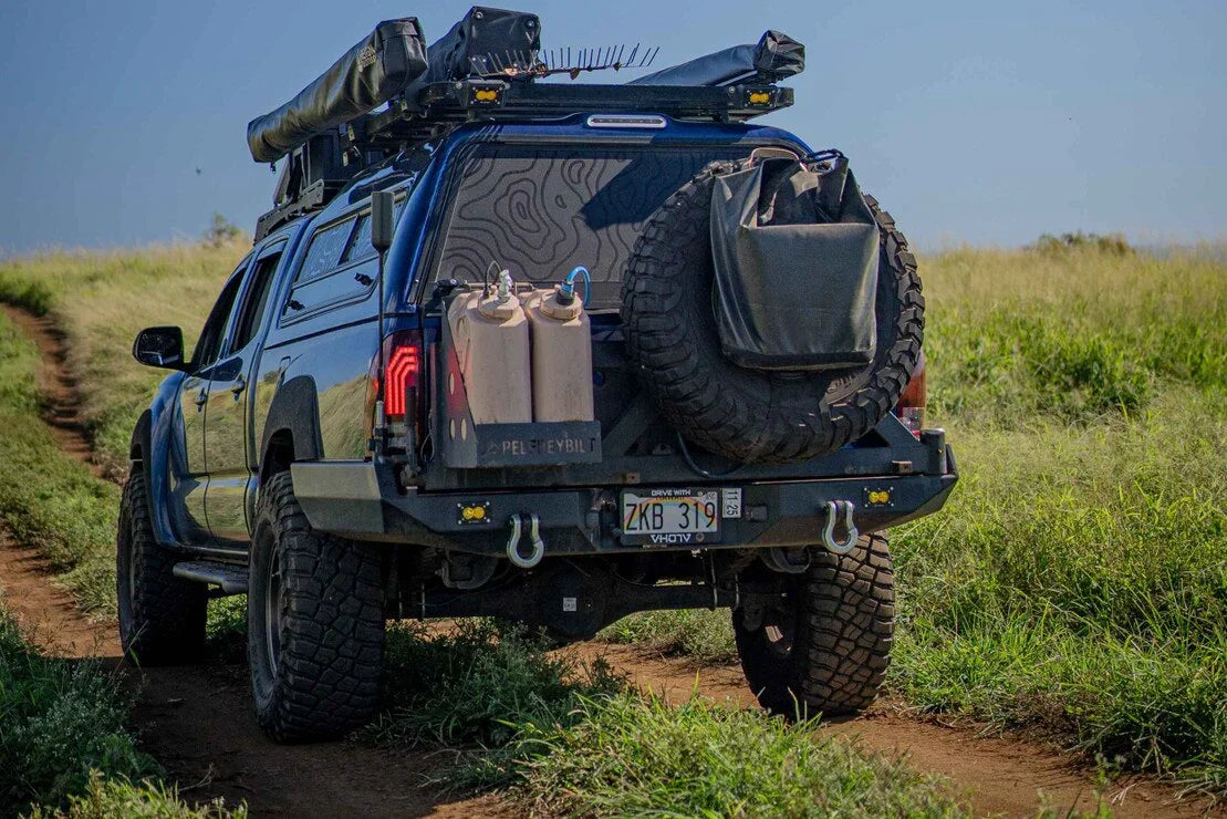 Off-road vehicle with camping gear, spare tire, and jerry cans on a dirt trail in grassy field