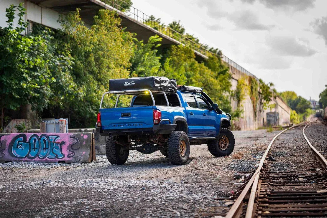 Blue lifted Toyota Tacoma with rooftop tent parked near railroad tracks and graffiti wall, overlanding setup.