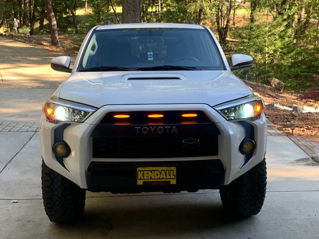 Front view of white Toyota 4Runner with amber grille lights and off-road tires in a wooded driveway
