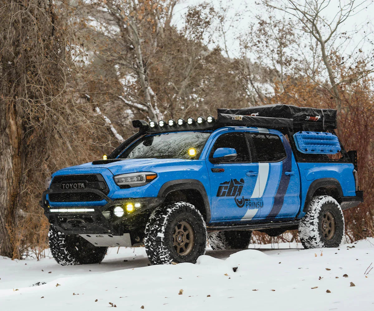 Lifted blue Toyota Tacoma with hybrid front bumper and overlanding gear on snowy trail