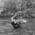 Man kneeling in river holding large fish, surrounded by trees, outdoor adventure scene