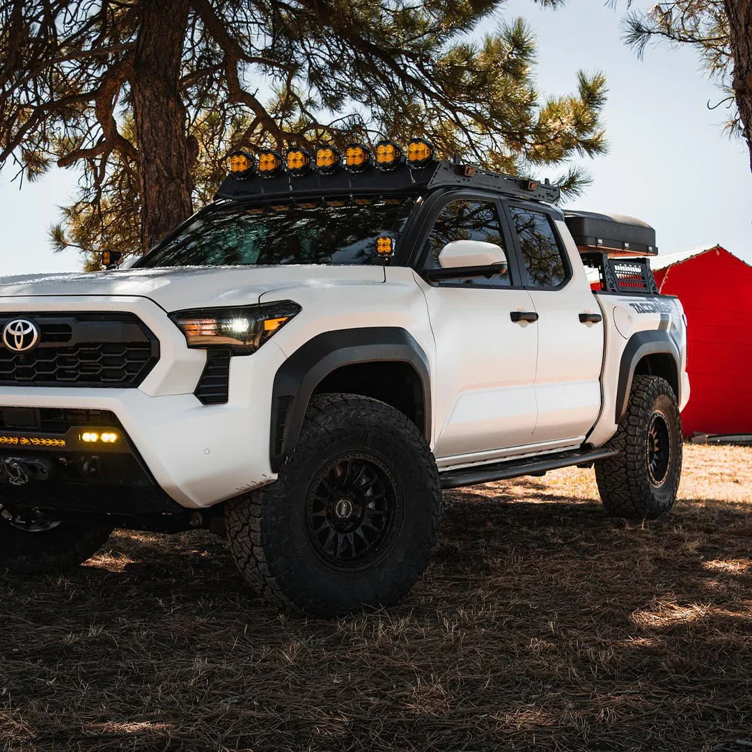 White Toyota Tacoma with off-road rock sliders, roof rack, and accessory lights parked outdoors