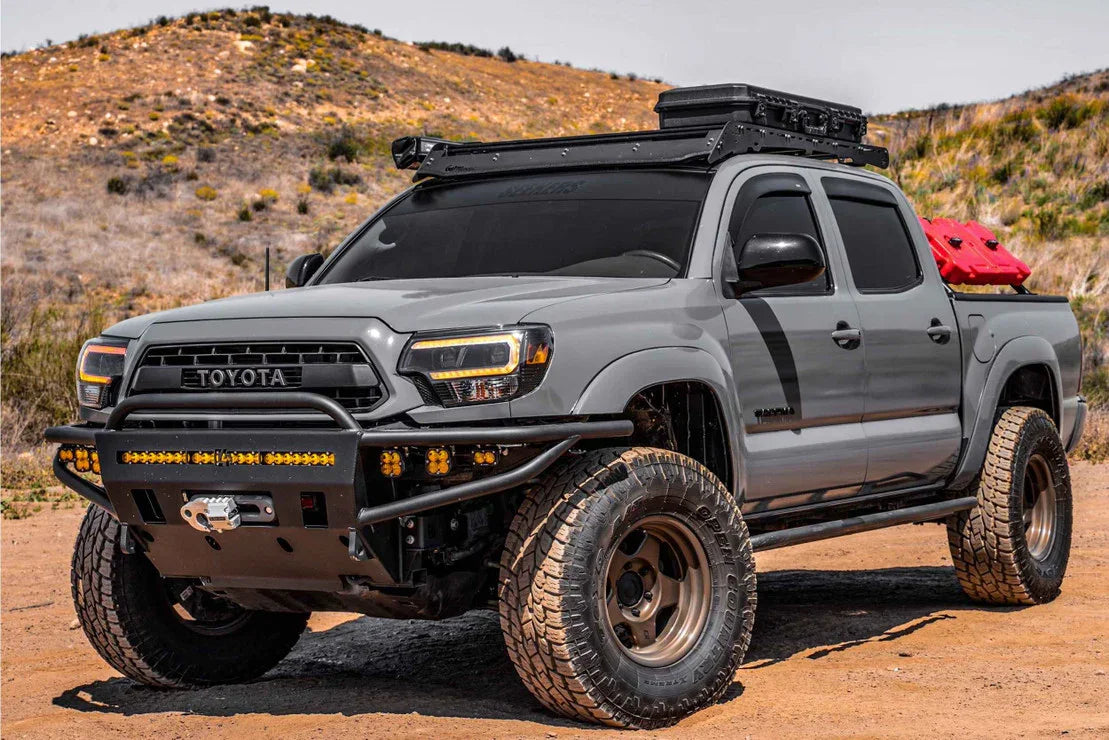 Modified gray Toyota Tacoma with off-road bumpers, amber hybrid headlights, roof rack, and gear, parked on a rugged trail.