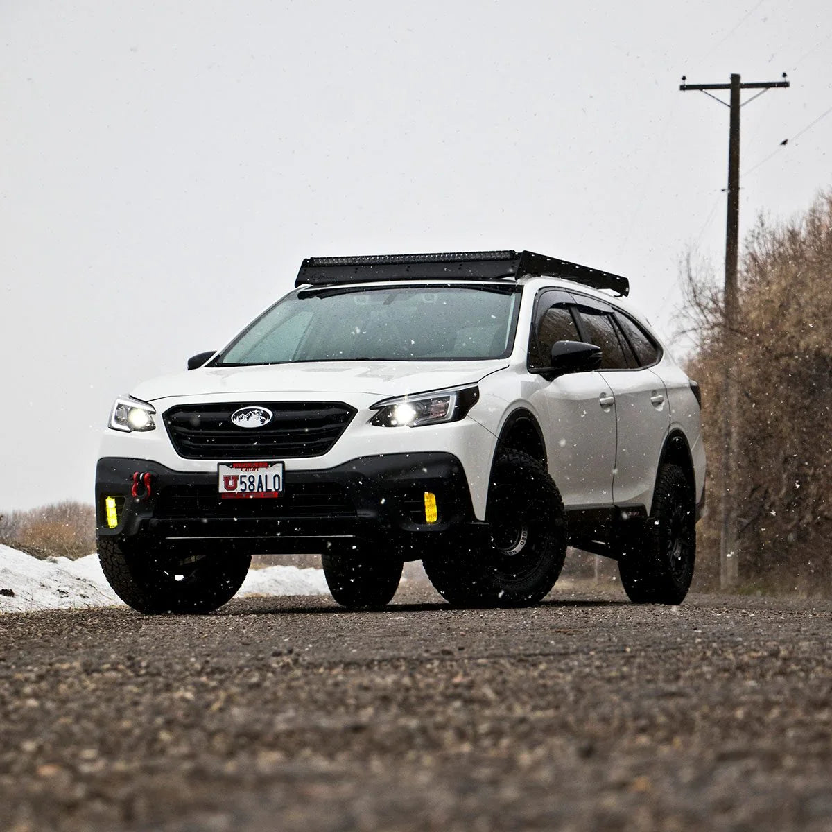 Lifted white Subaru Outback with off-road tires and roof rack, parked on gravel road in winter.