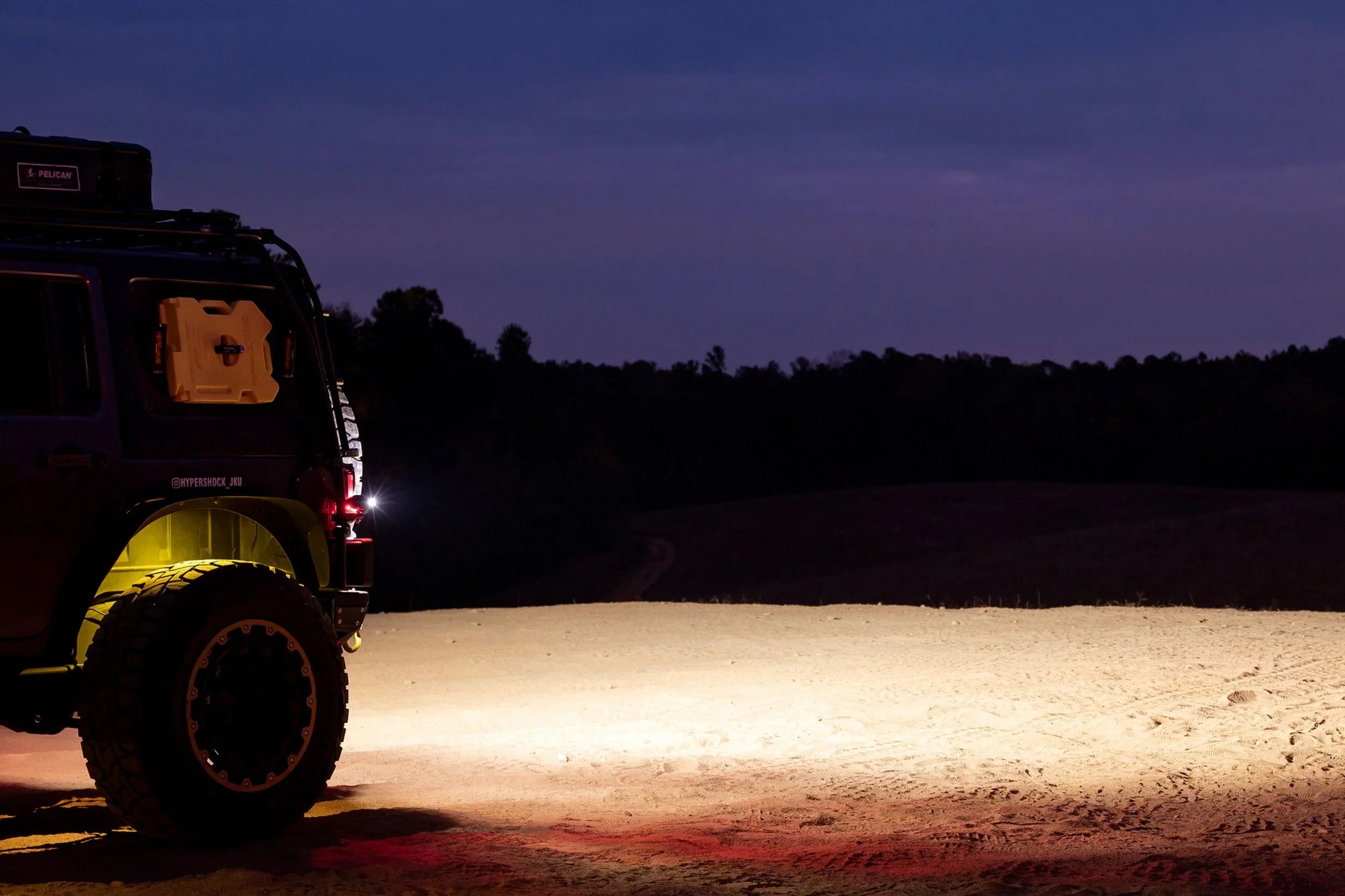 Off-road vehicle with large tires and roof gear at dusk, overlanding on a sandy trail