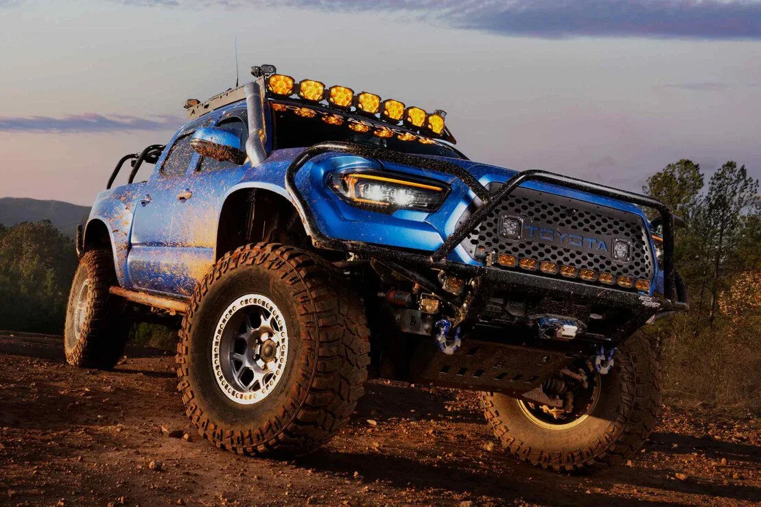 Blue Toyota Tacoma outfitted with off-road lights, rugged bumper, and mud-terrain tires on a dirt trail