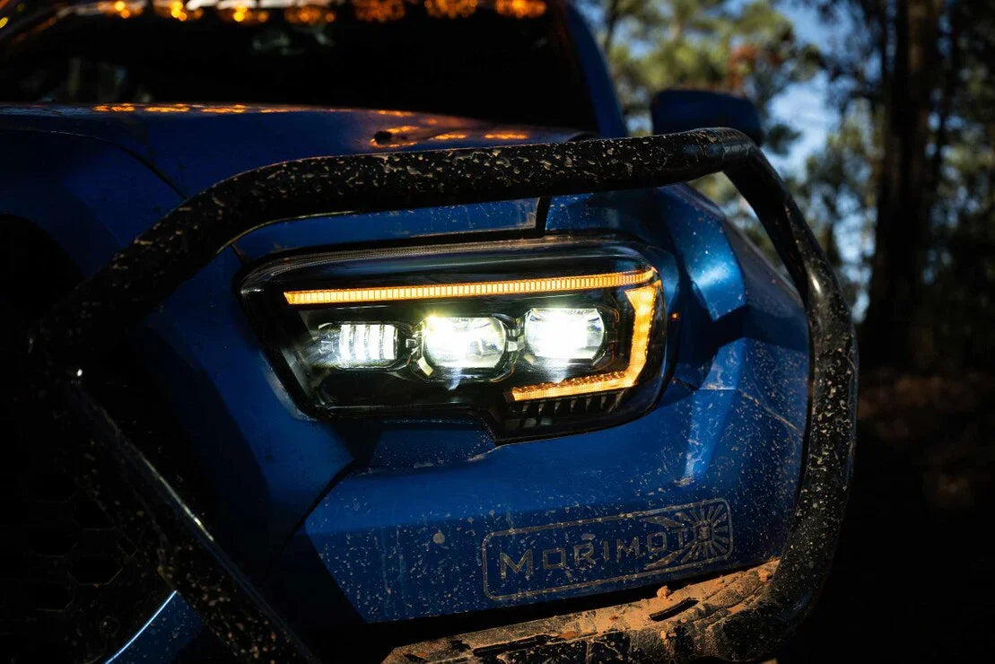 Close-up of LED projector headlight on a blue Toyota Tacoma off-road vehicle with mud splatters.