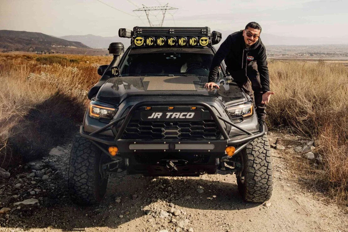 Lifted Toyota Tacoma with off-road lights, rugged bumpers, and a man on the hood on a dirt trail