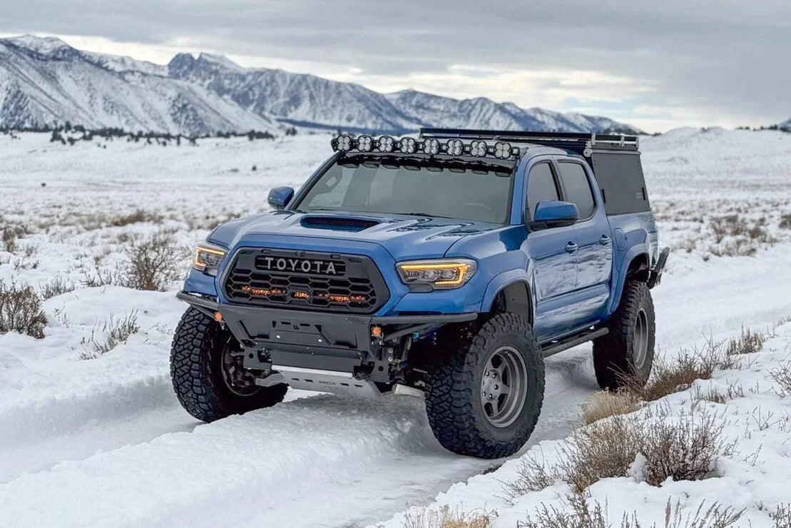 Blue Toyota Tacoma overland truck with off-road lights in a snowy mountain landscape