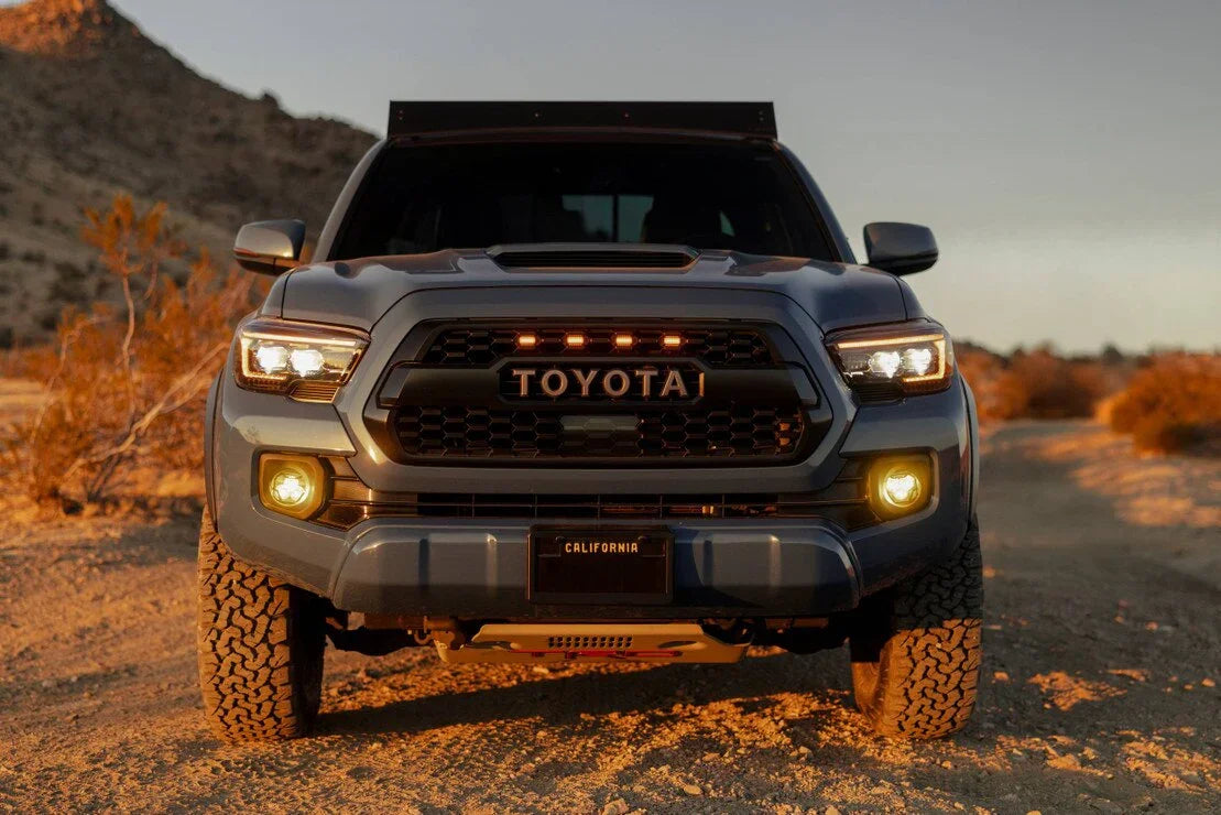 Toyota Tacoma with aftermarket headlights and grille lights on a desert trail at sunset