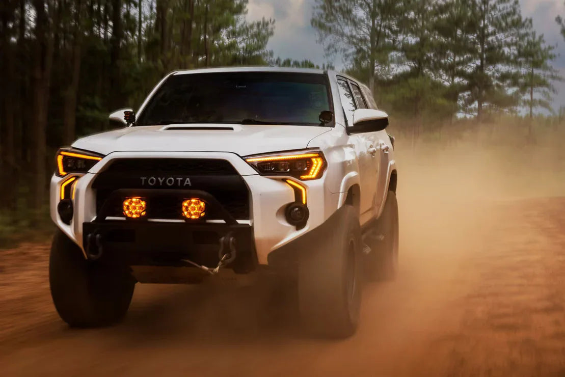 White Toyota 4Runner with off-road lights driving on a dirt trail in the forest