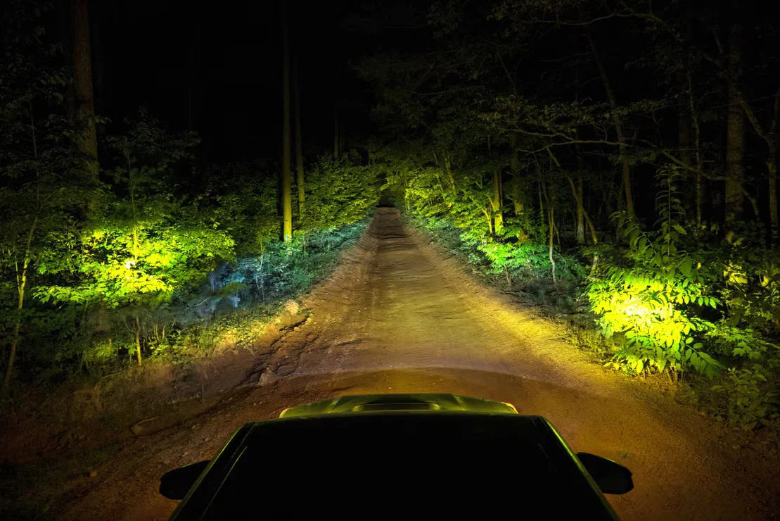 Off-road vehicle driving at night on a dirt trail illuminated by bright yellow auxiliary lights.