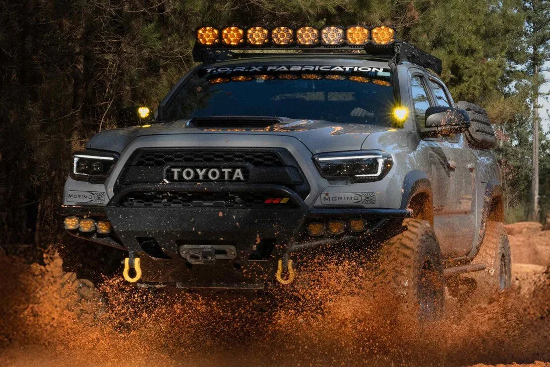 Toyota Tacoma off-road truck with upgraded Morimoto headlights and amber LED light bar driving through mud