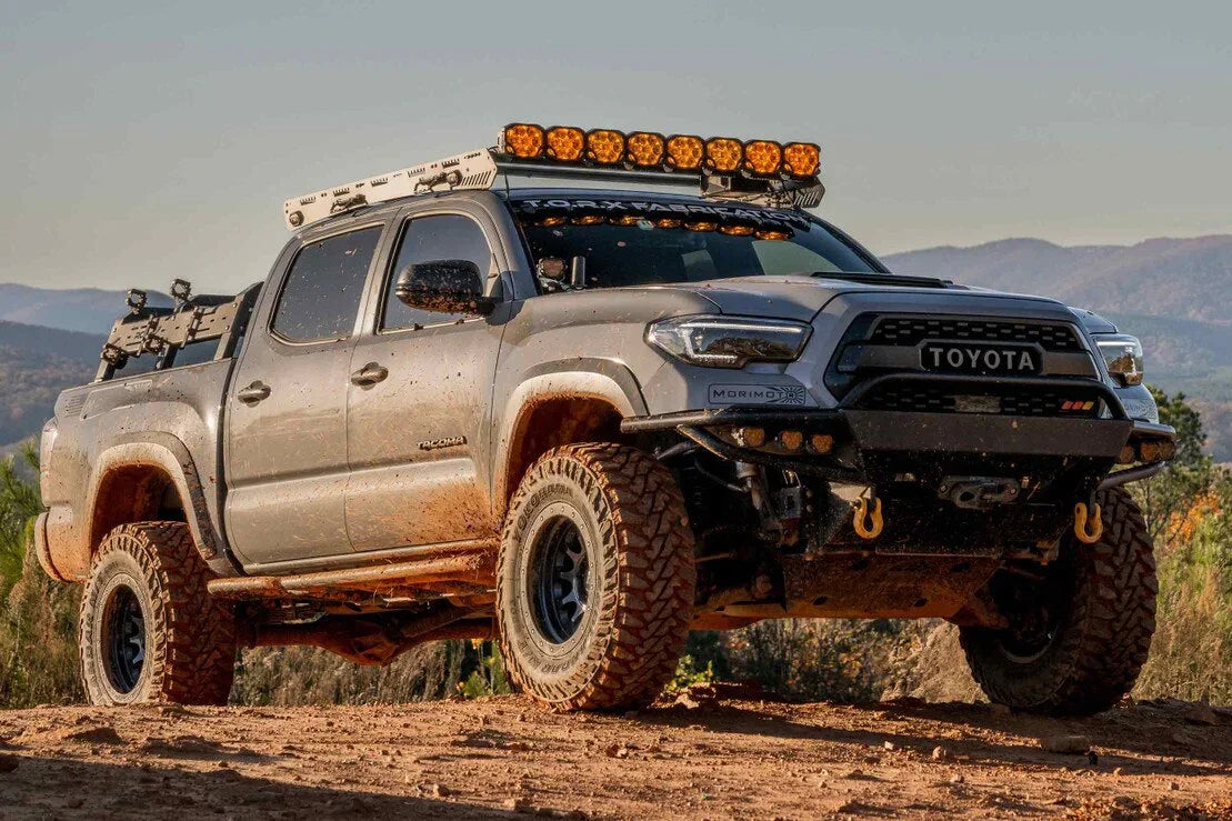Lifted Toyota Tacoma off-road truck with aftermarket headlights, roof lights, and rugged tires on a dirt trail