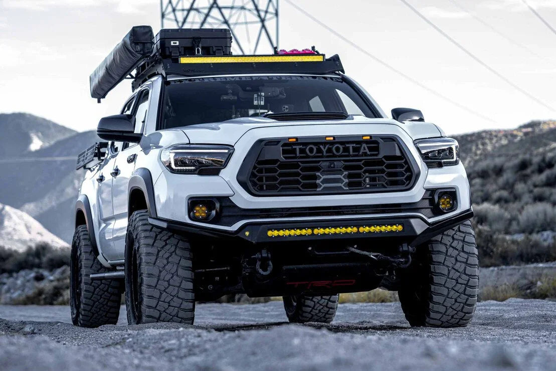 Lifted white Toyota Tacoma with aftermarket hybrid headlights, off-road lights, and roof rack in rugged mountain terrain
