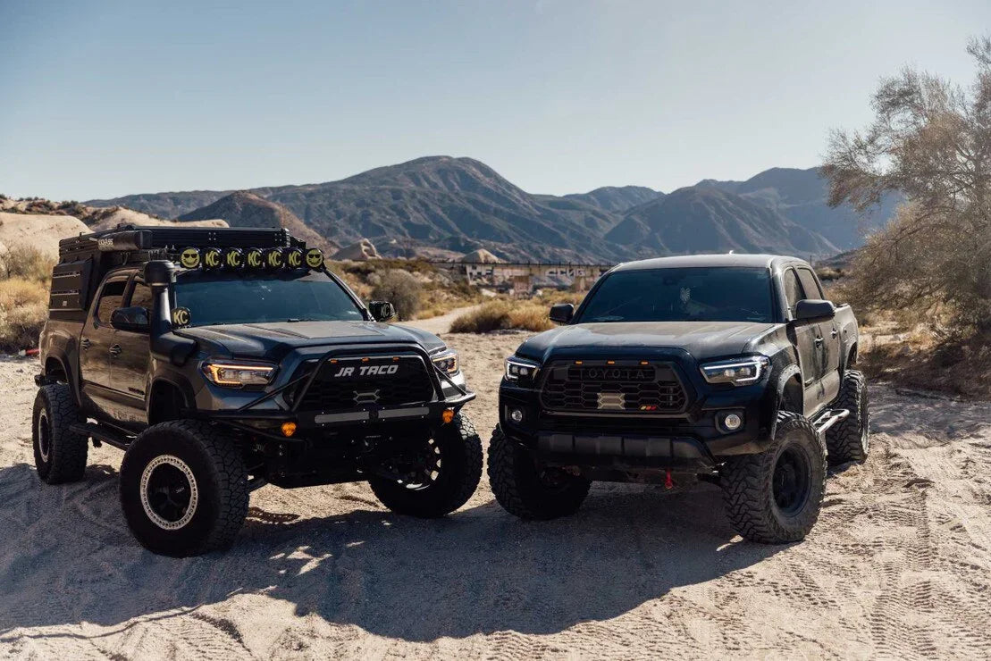 Two lifted Toyota Tacoma trucks with off-road accessories on a desert trail, mountains in background.
