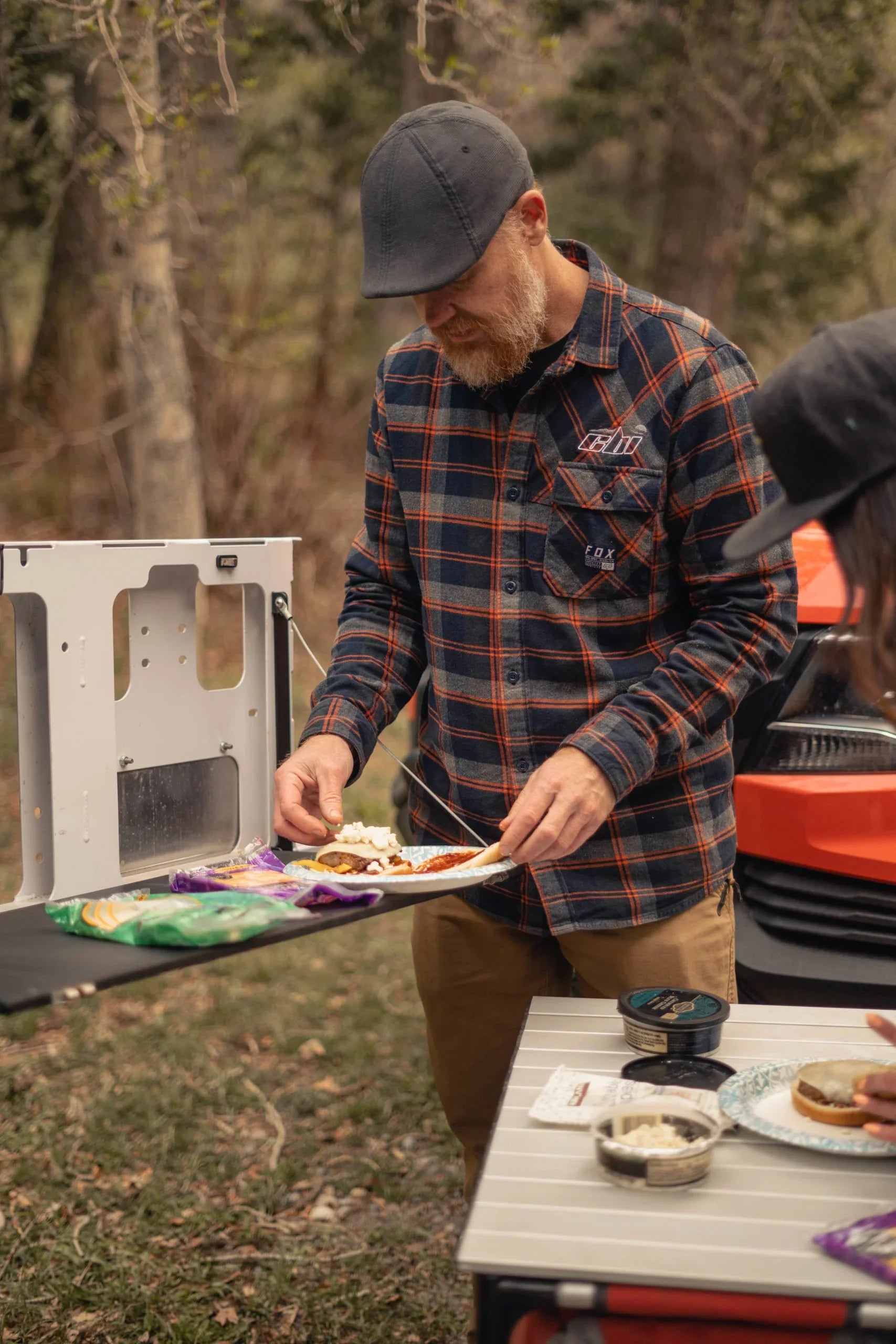 Man using swing arm camp table accessory for outdoor cooking near vehicle in forest