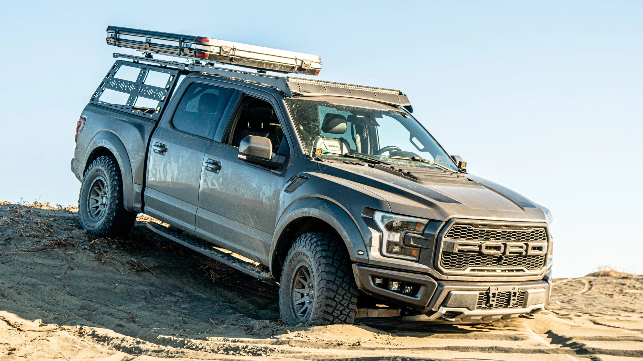 Ford Raptor truck with off-road bed rack and roof gear driving on sandy terrain