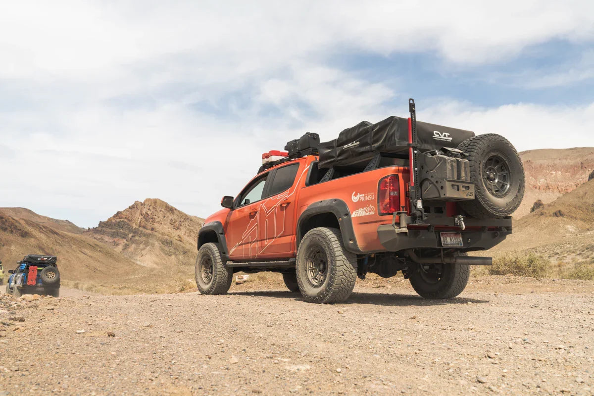 Red overland truck equipped with camping gear, roof rack, and spare tire in desert terrain