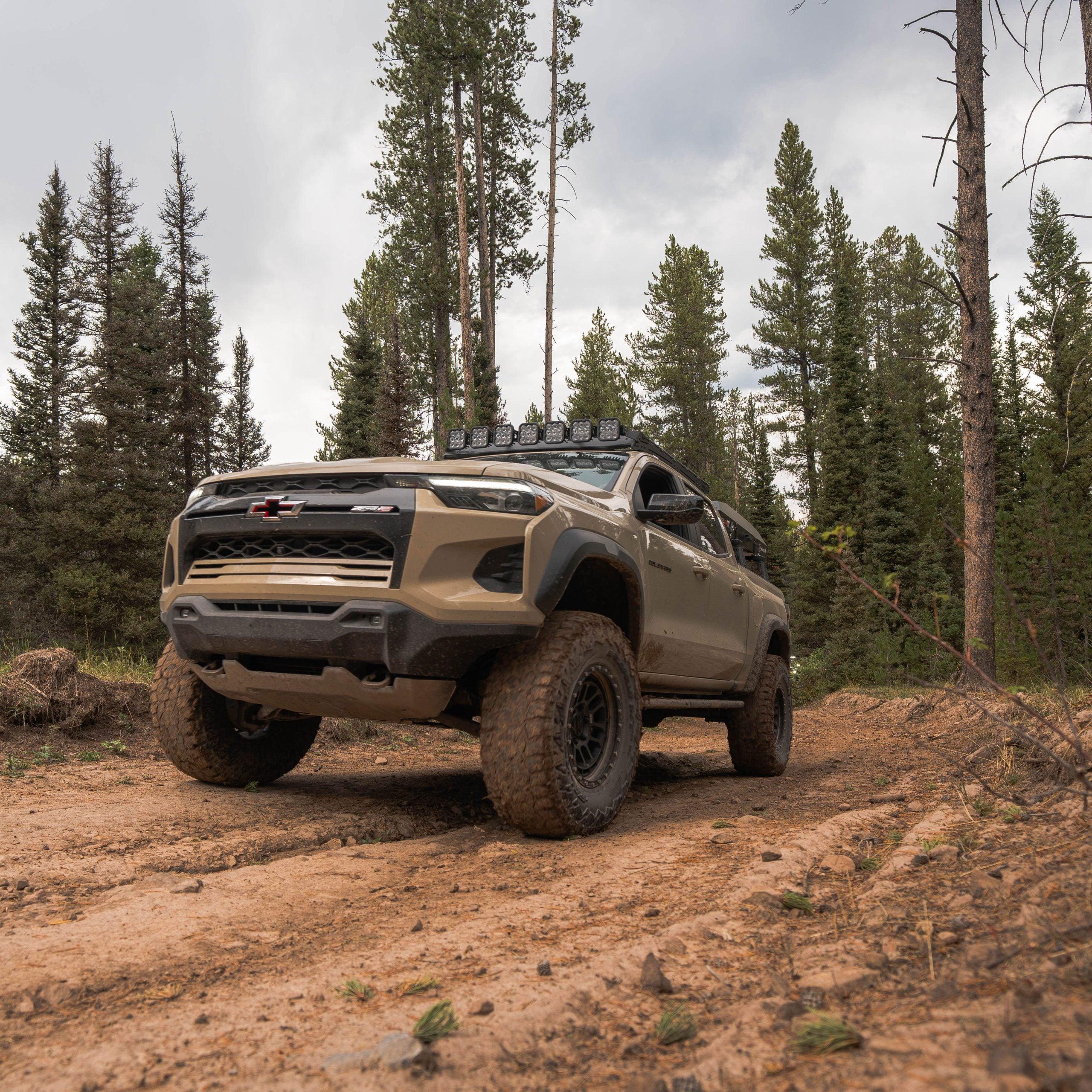 Lifted off-road truck with light bar driving on a forest dirt trail, overlanding gear visible