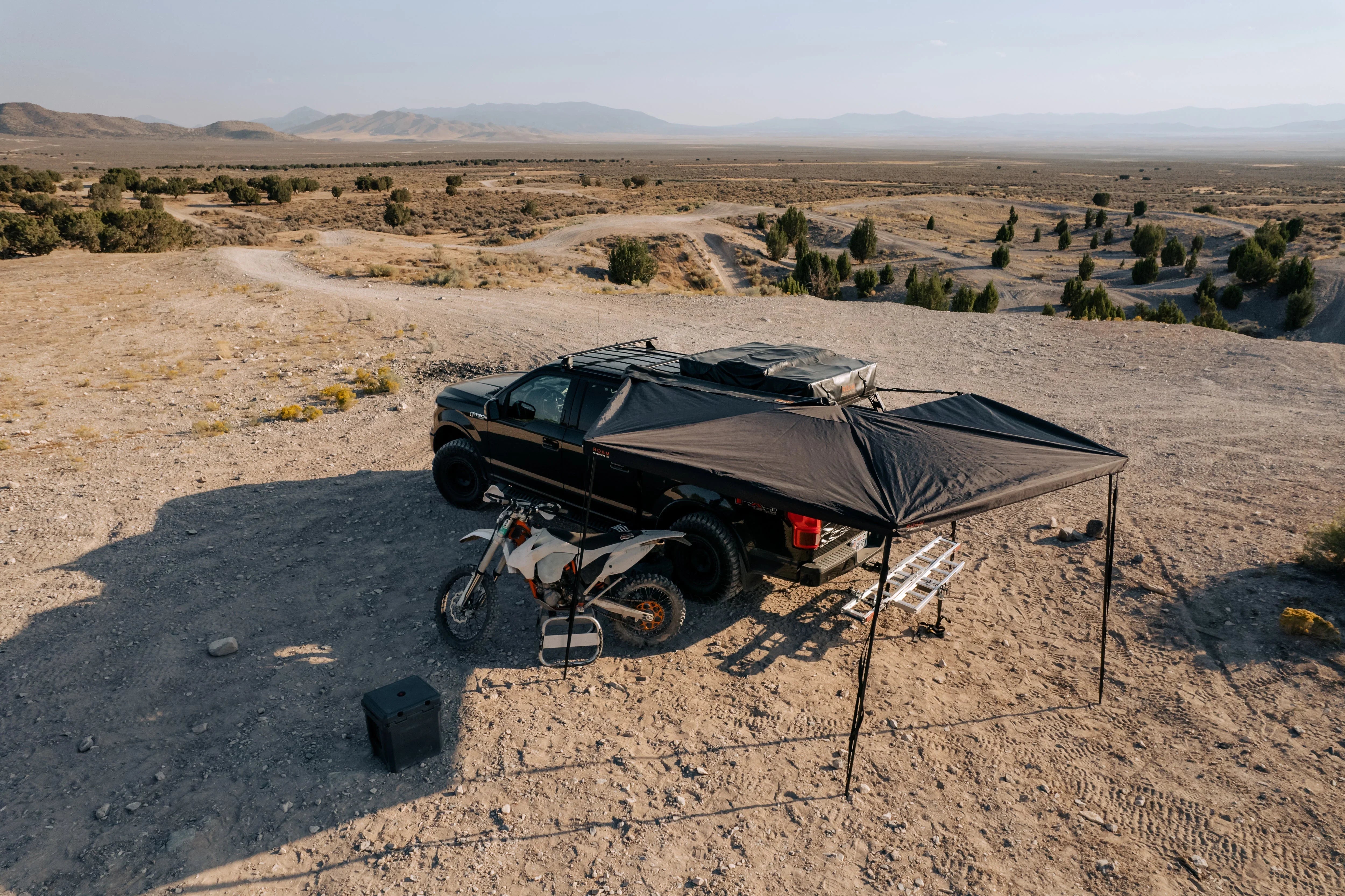 Overland truck with rooftop tent, awning, and dirt bike set up for camping in a desert landscape