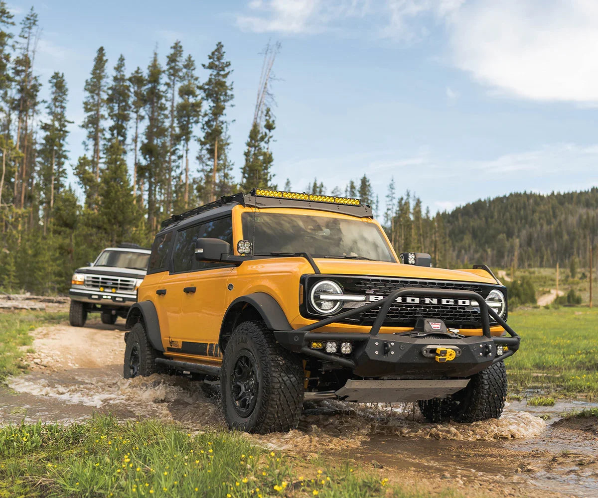 Yellow Ford Bronco with bolt-on rock sliders off-roading through mud in forest terrain