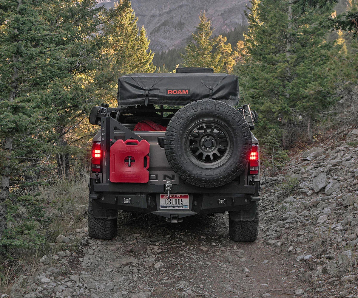 Overland truck with spare tire, red fuel can, and rooftop tent on rocky forest trail