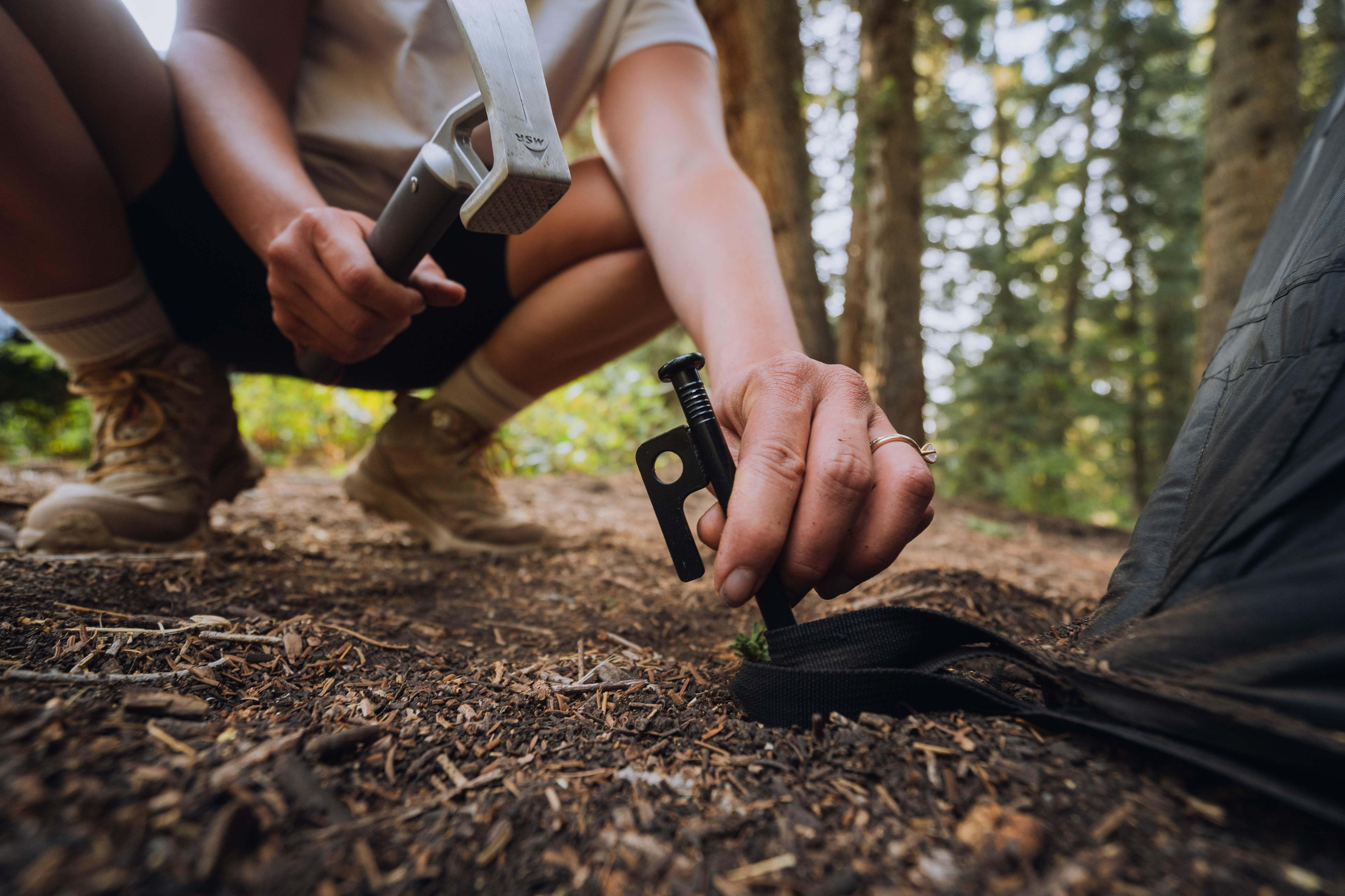 Person staking down a tent in a forest, using camping gear for outdoor adventure setup