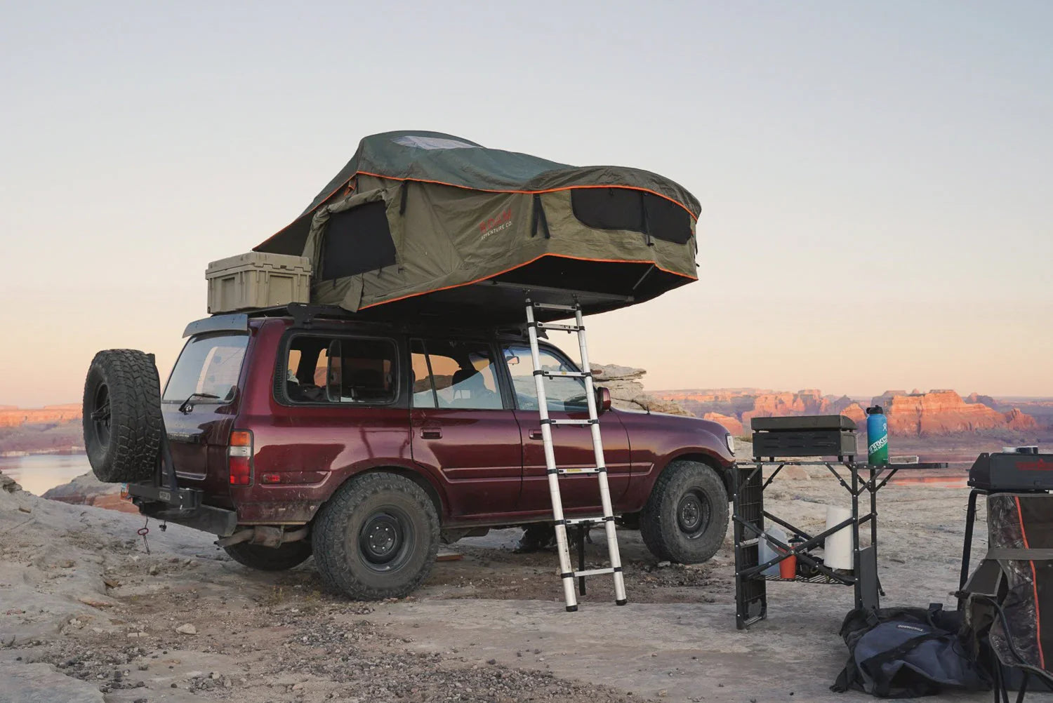 SUV with rooftop tent and camping gear set up on rocky overlook at sunset, overlanding scene
