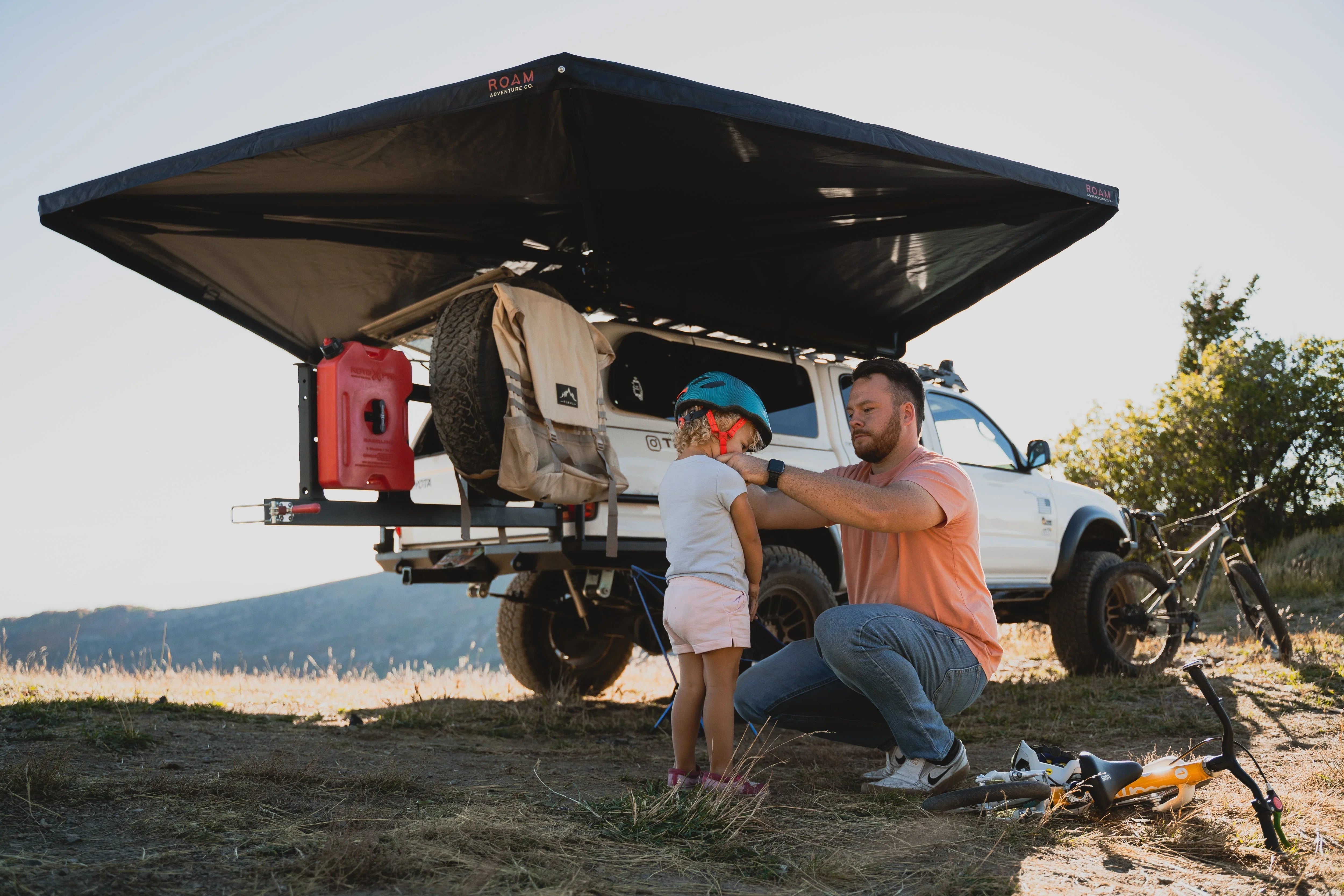 Man helping child with helmet near overlanding Toyota truck, camping awning, and bikes outdoors