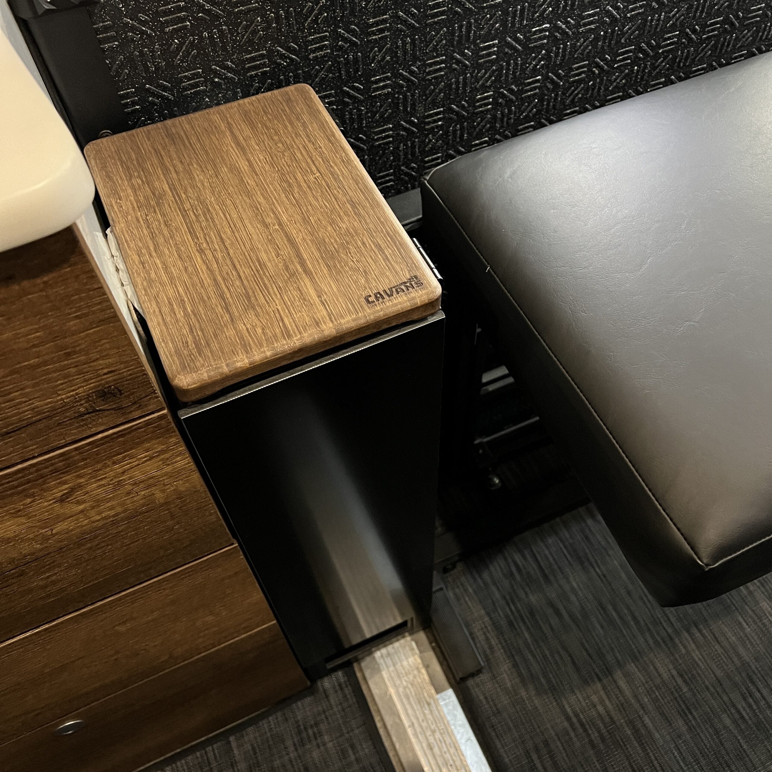 Compact camper van interior with wood and metal storage compartment, black seat, and textured flooring