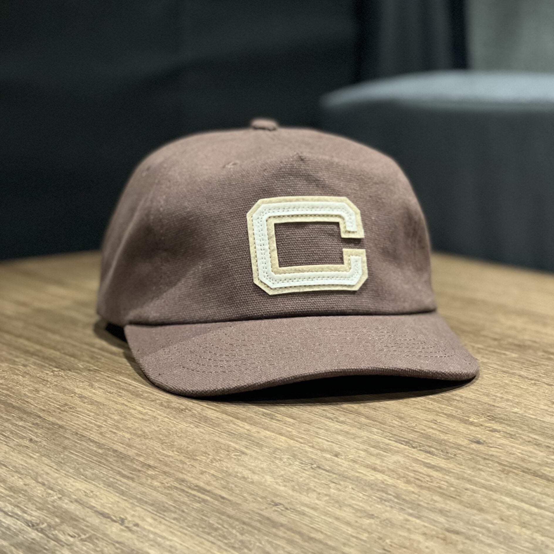 Brown cotton baseball cap with a large embroidered C on the front, on wooden table