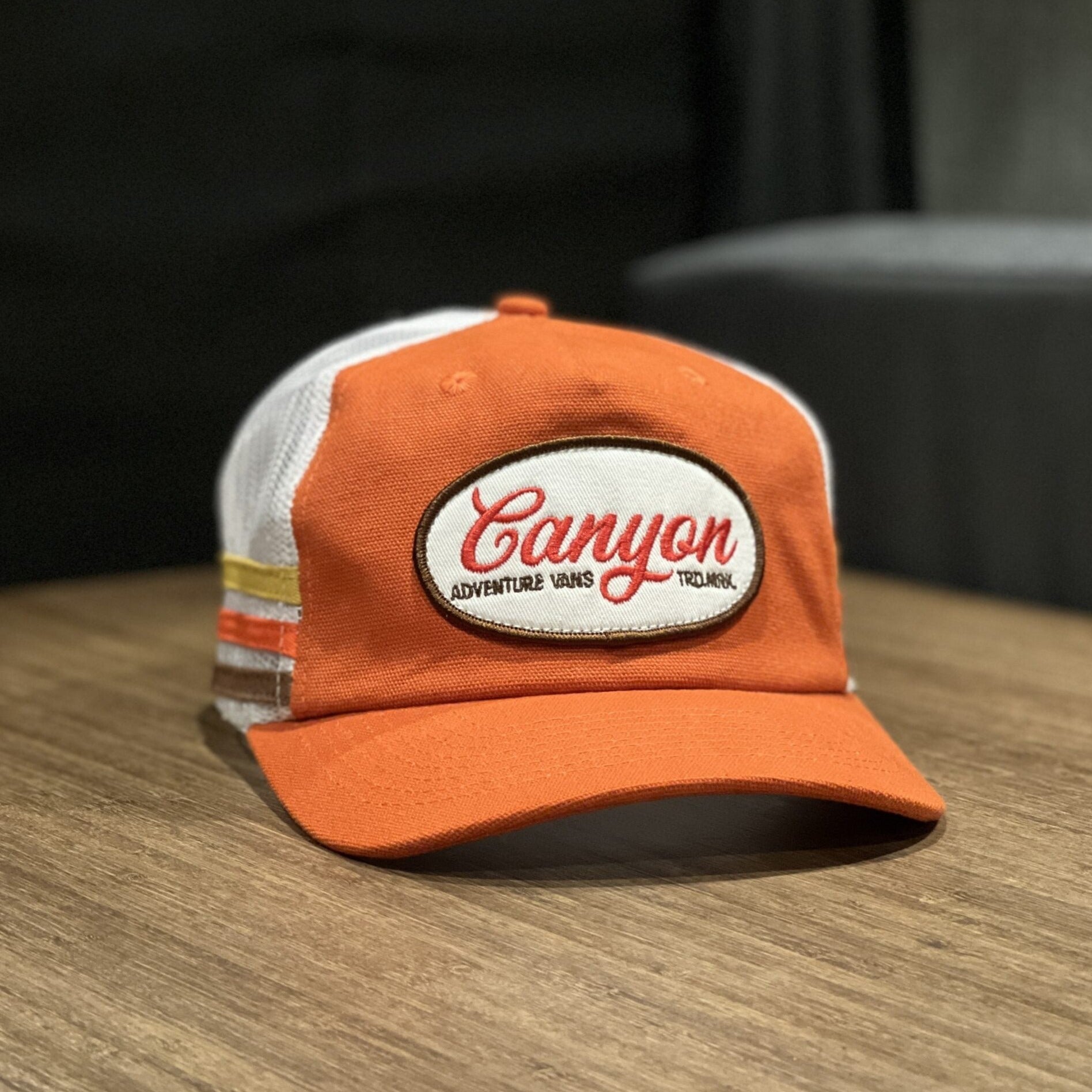 Orange and white trucker cap with 'Canyon Adventure Vans' patch on wooden table