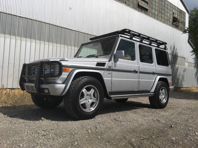 Silver off-road SUV with roof rack and bull bar parked on gravel in front of industrial building