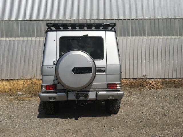 Rear view of a silver off-road SUV with roof rack and spare tire, parked on gravel