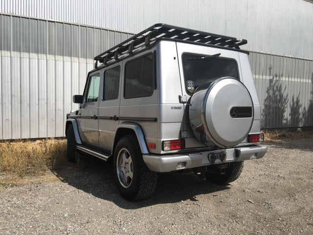 Silver Mercedes G-Class SUV with roof rack parked outdoors on gravel by metal building