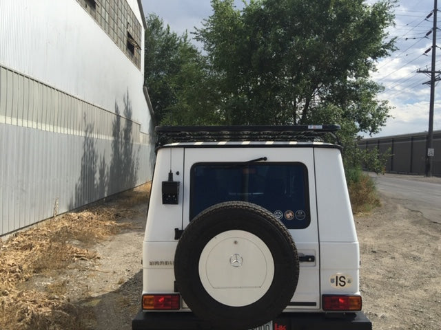 Rear view of a white Mercedes G-Wagon with roof rack and spare tire, parked by a warehouse