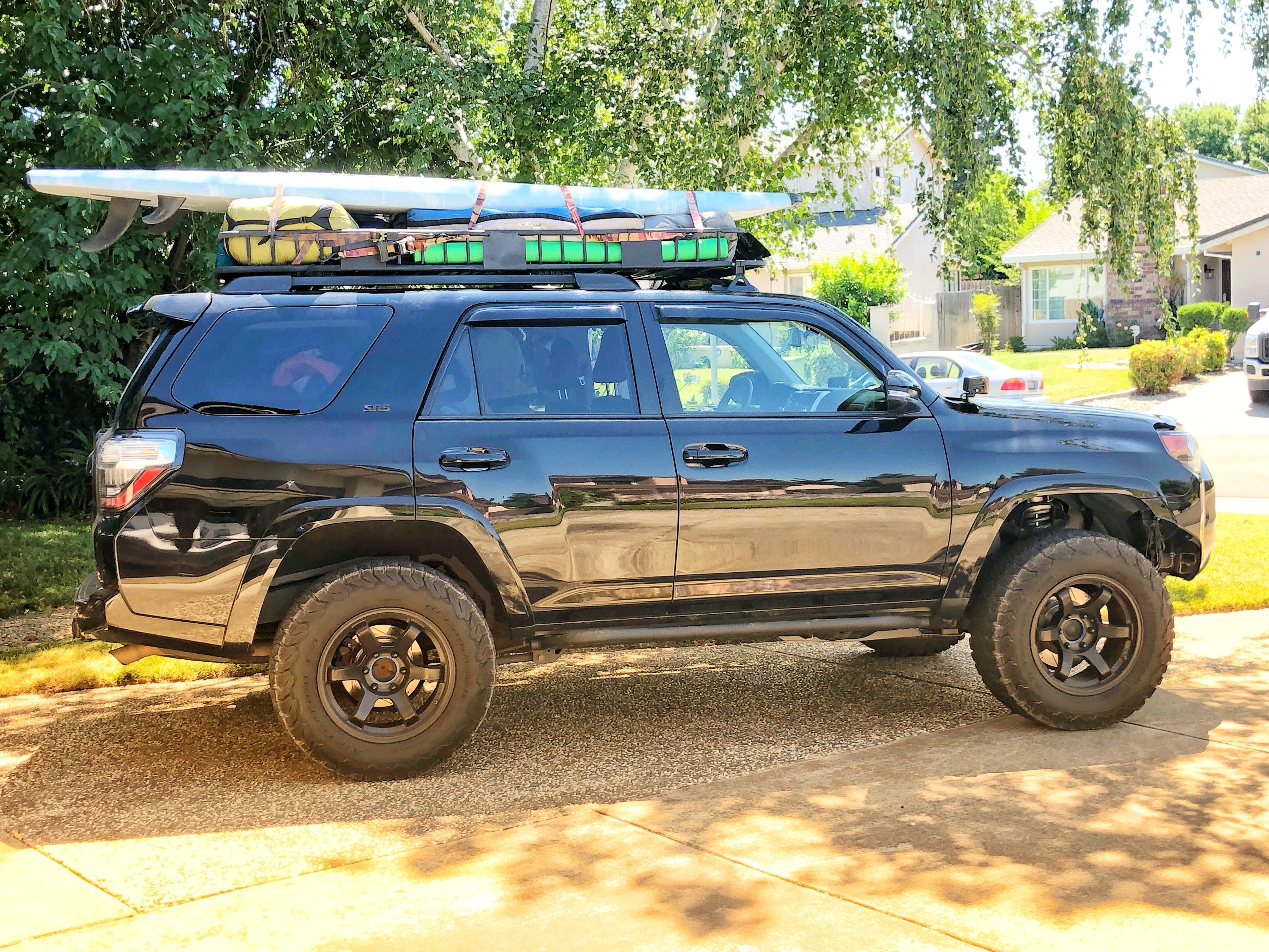 Black Toyota 4Runner with roof rack carrying camping gear and surfboard, parked outdoors.
