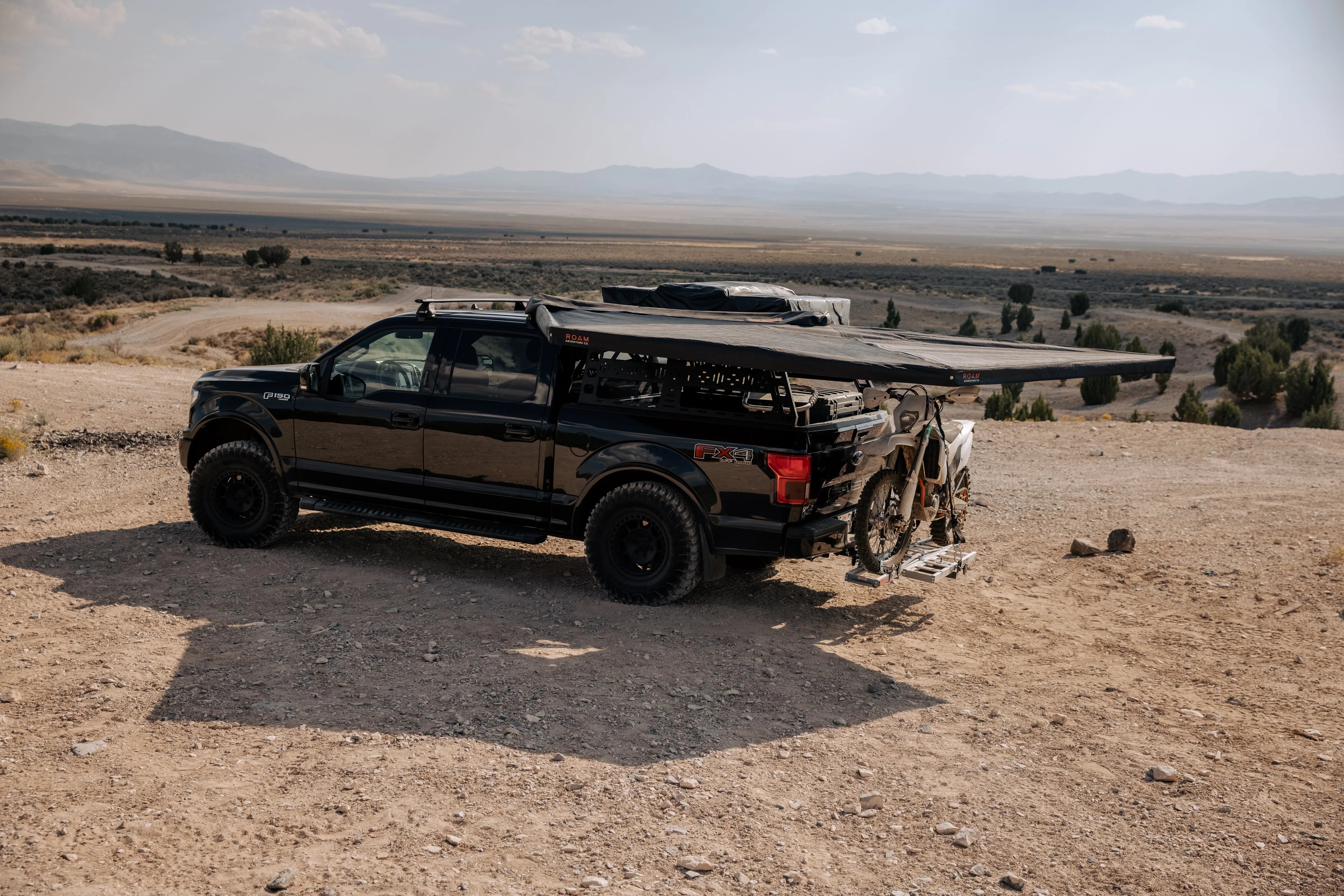 Off-road truck with rooftop tent, awning, and dirt bike in desert landscape