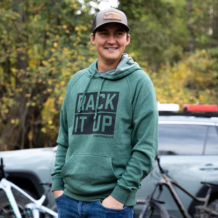 Man in green 'Rack It Up' hoodie and cap outdoors near overland vehicle and bicycle