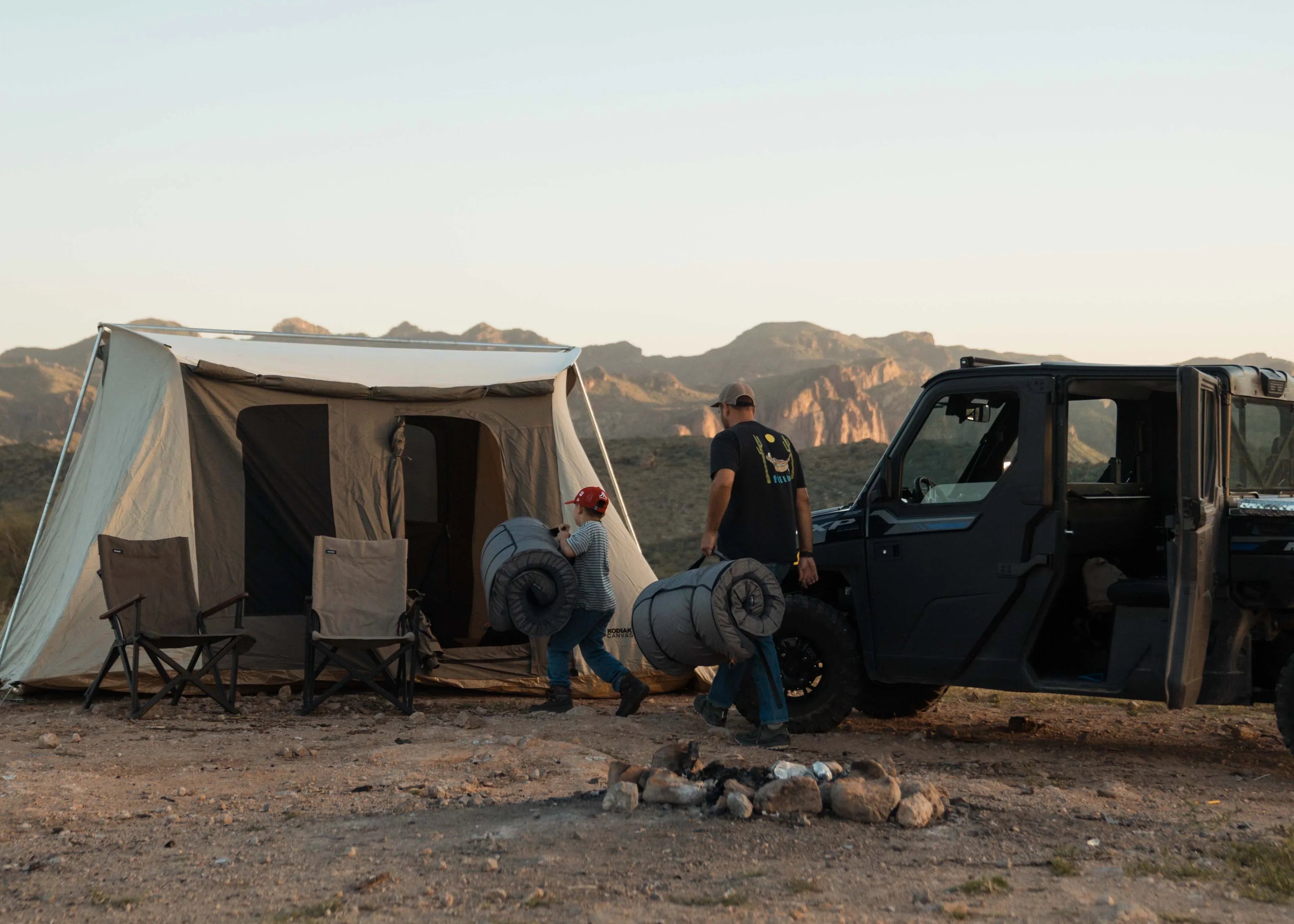 Father and son carry camping mats to tent beside off-road vehicle in rugged outdoor setting