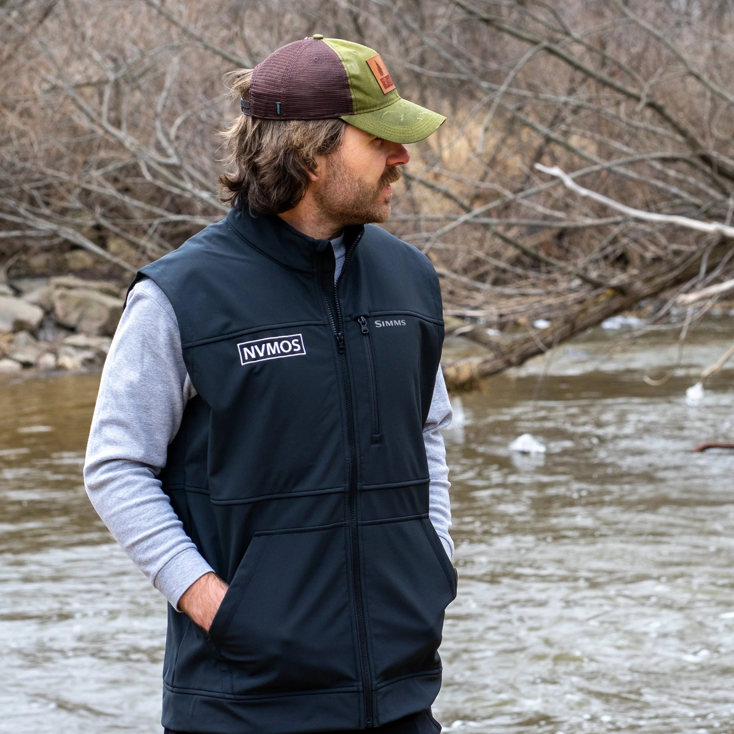Man wearing black outdoor vest and cap standing by river in adventure gear setting
