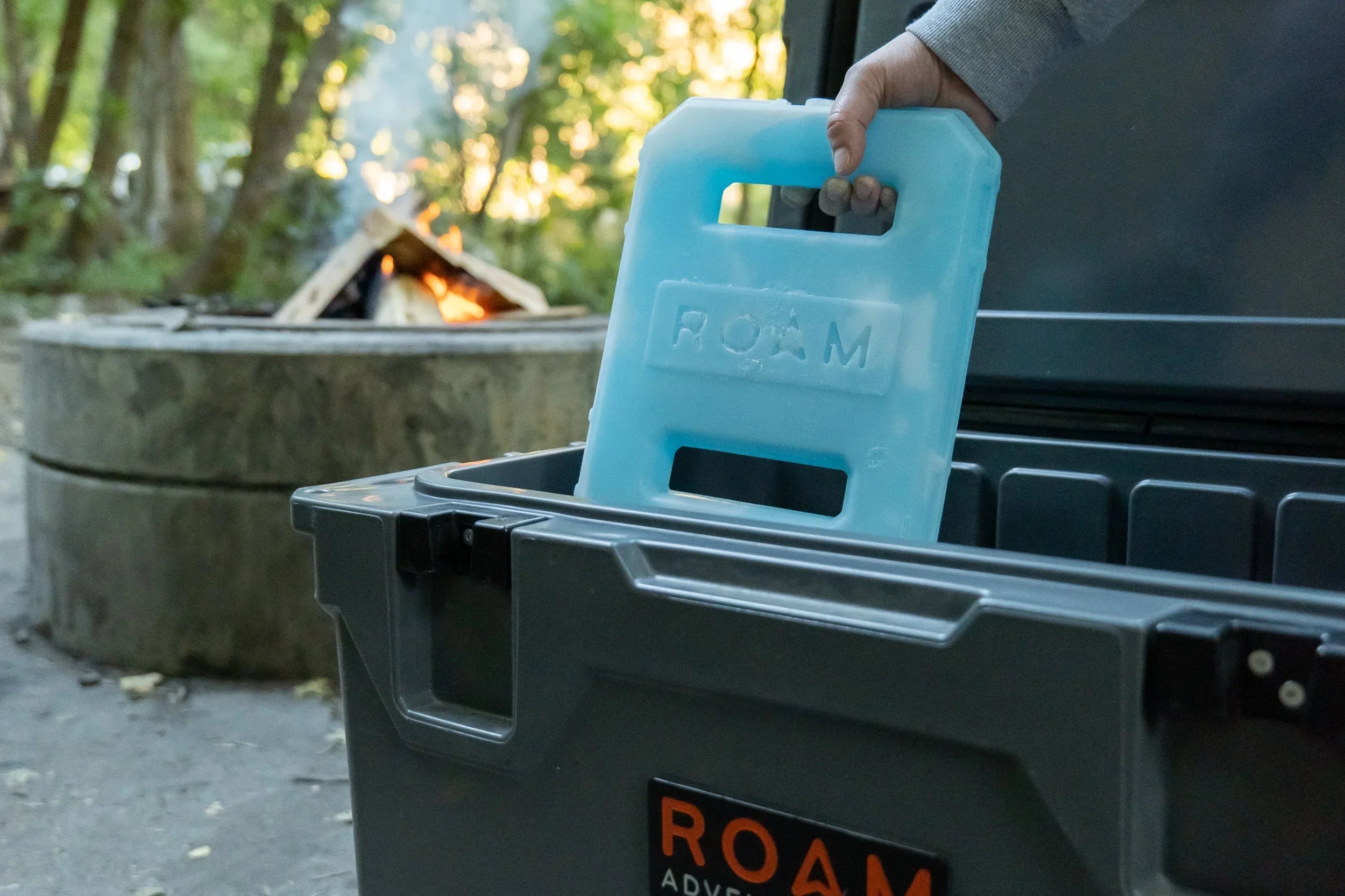 Camping ice pack being placed in a storage box near campfire outdoors, overlanding gear setup.