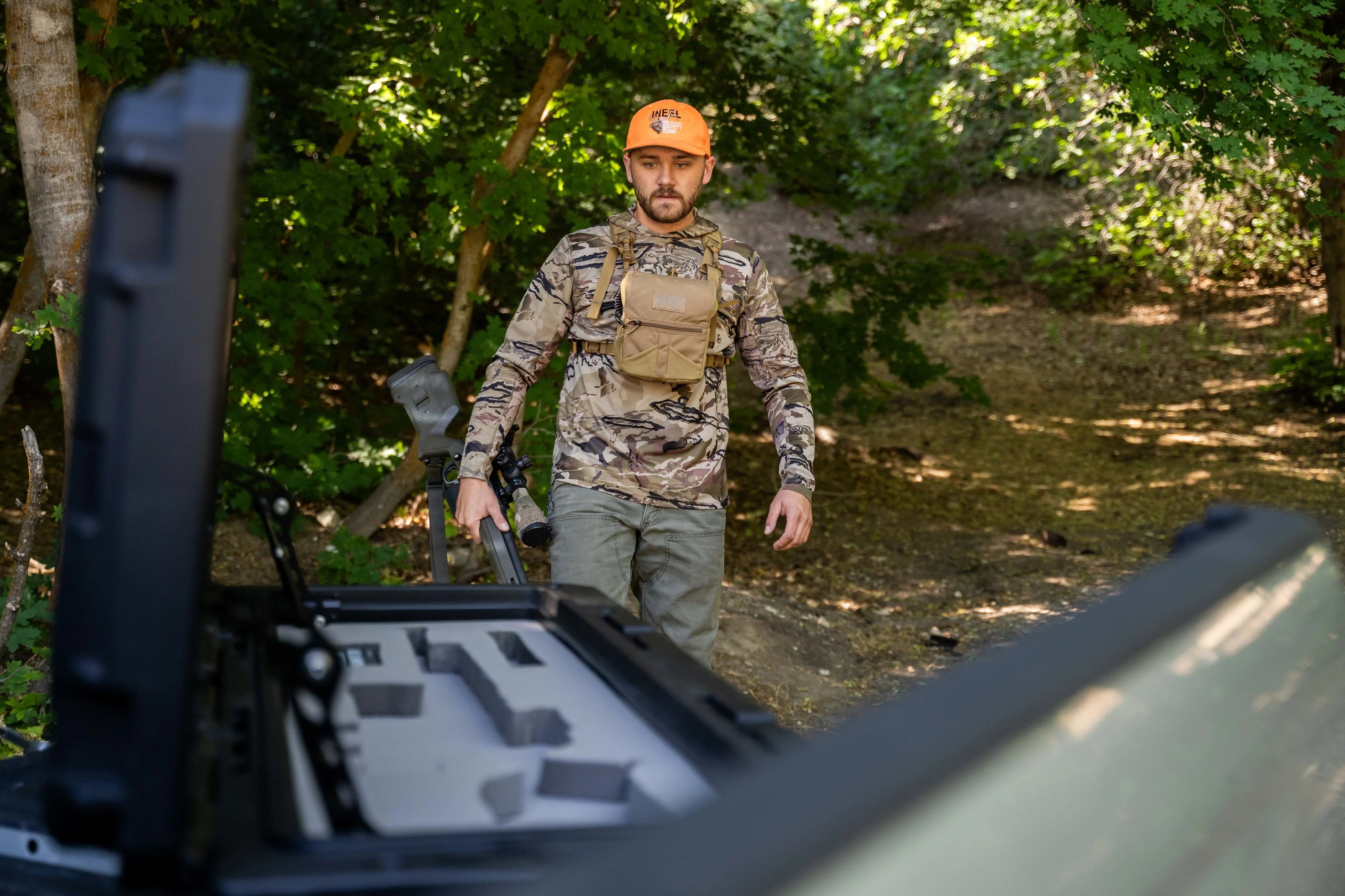 Man in camo gear with rifle walking toward open truck bed storage box in forest setting