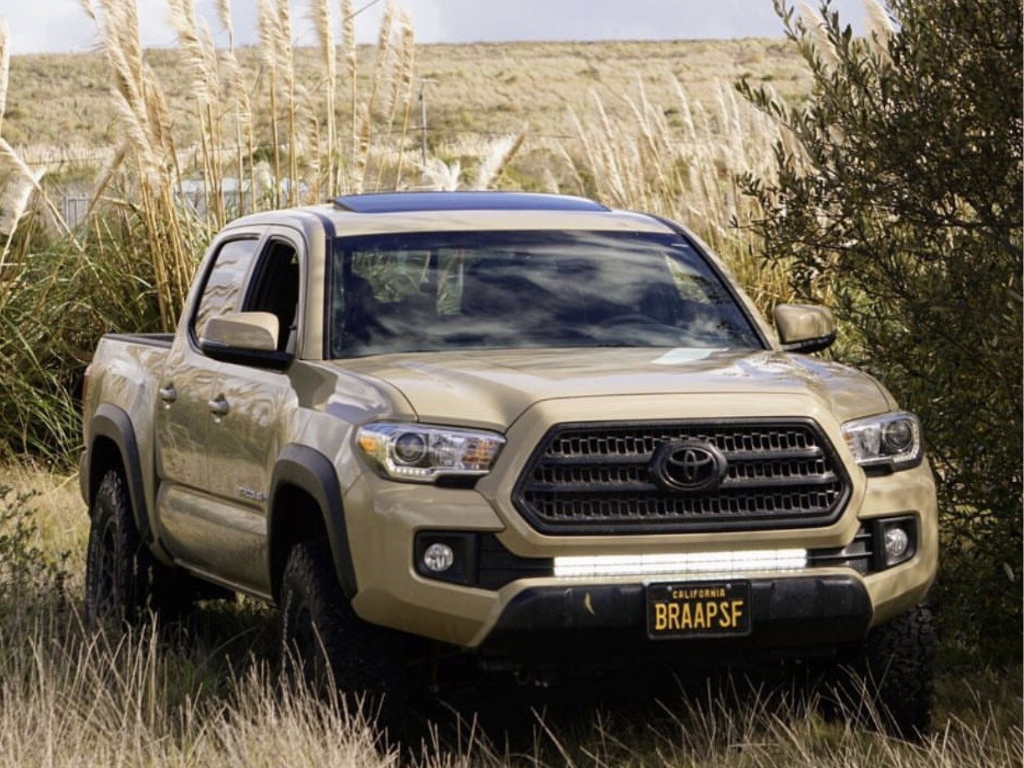 Beige Toyota Tacoma pickup truck with off-road tires and LED light bar in grassy outdoor setting