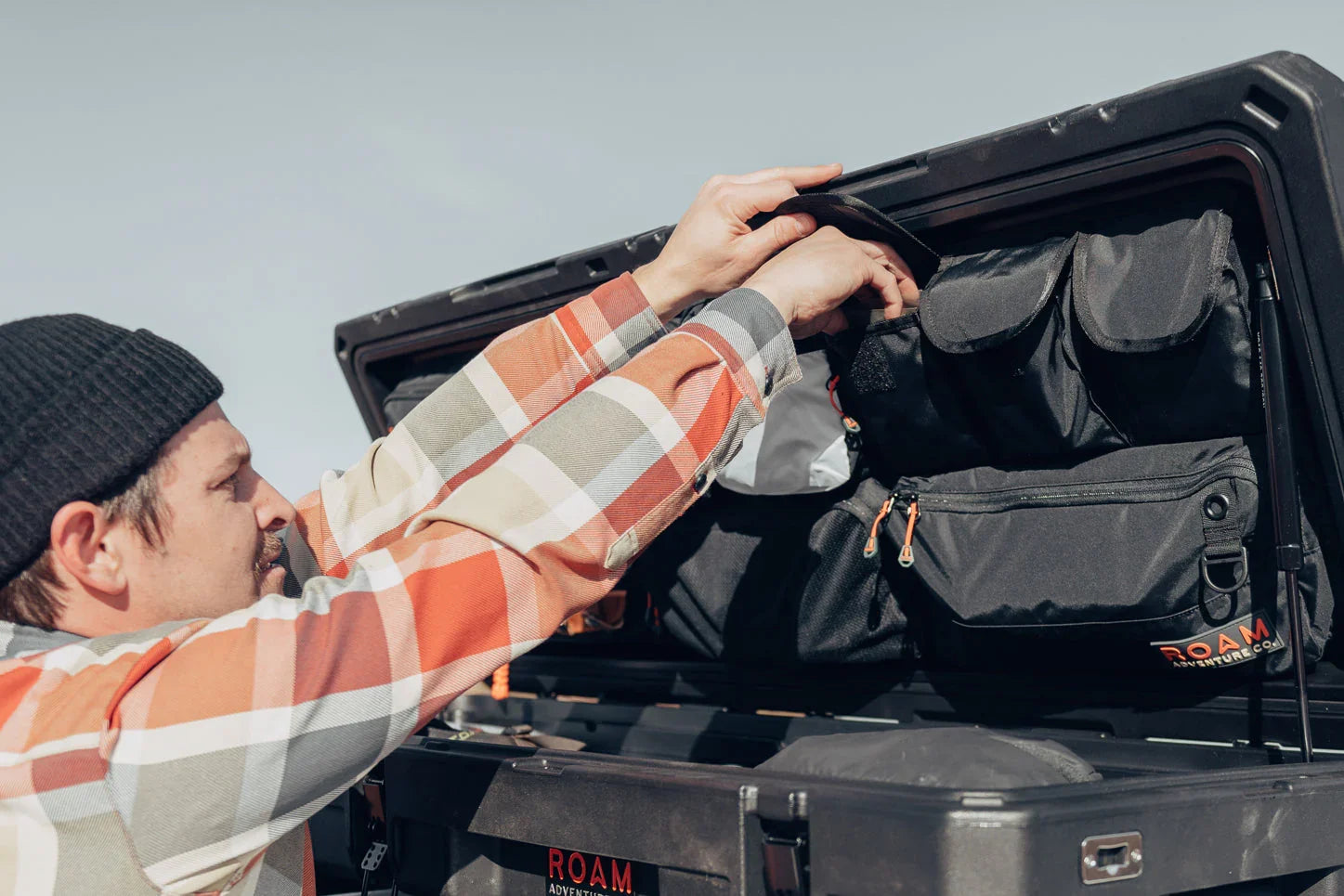 Man organizing black overland gear bags in vehicle rooftop storage box outdoors