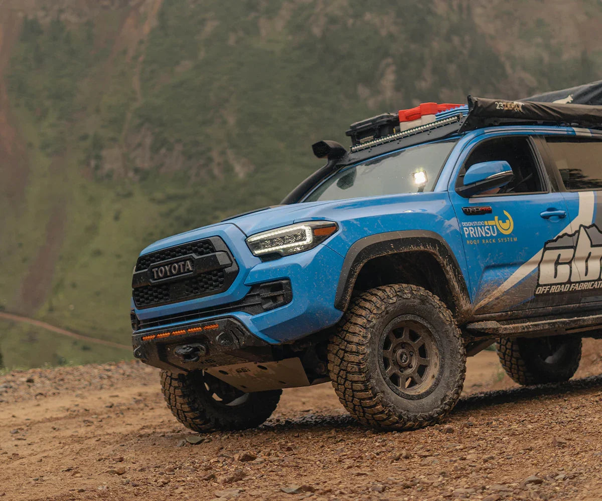 Blue Toyota Tacoma with Covert bumper, off-road tires, and roof rack on a dirt trail